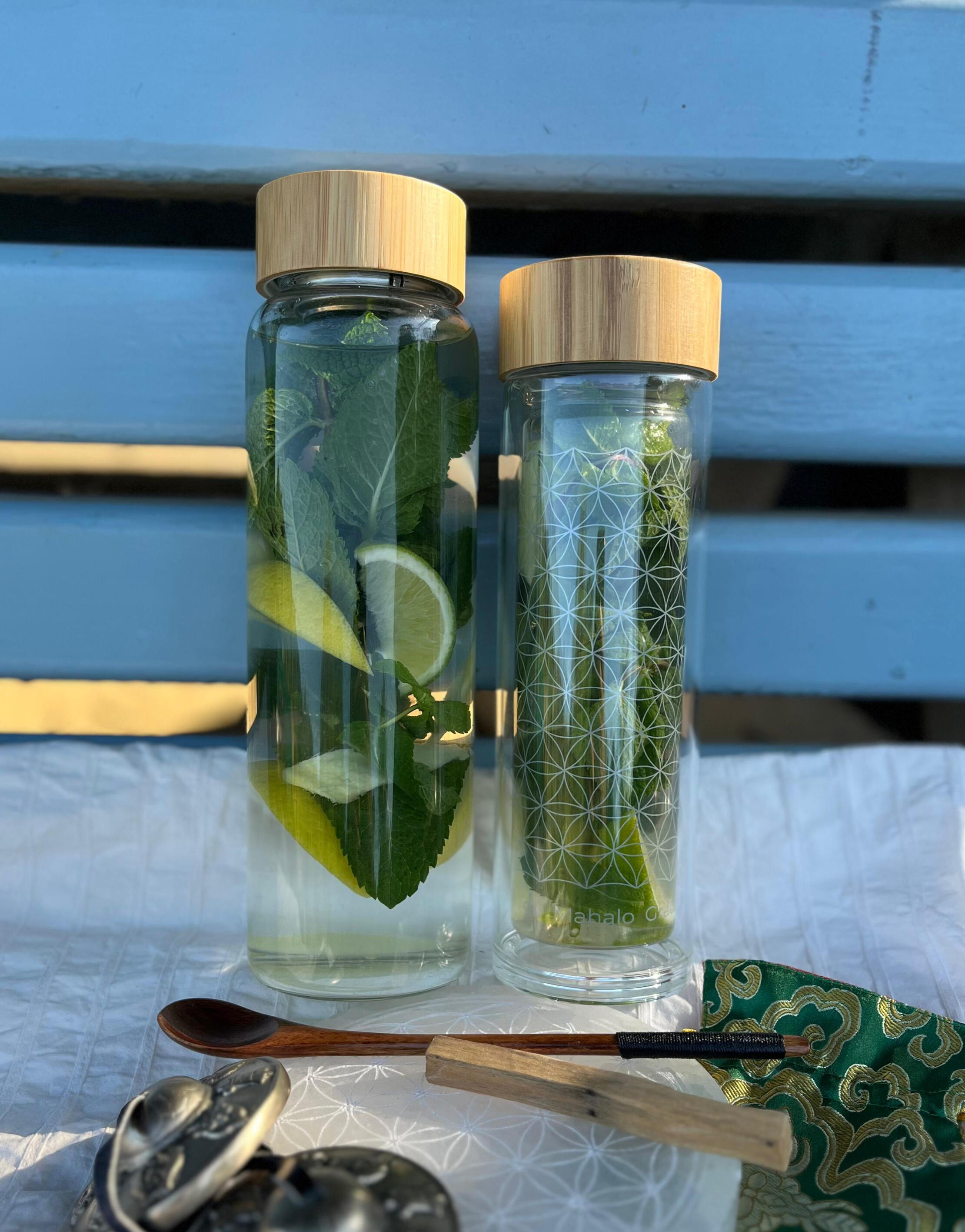 AMAZI Sacred Tea Bottles with Flower of Life Design Two AMAZI glass tea bottles with Flower of Life engraving on blue bench, filled with fresh herbs and lemon water, surrounded by ritual tools including wooden spoon and sacred geometry cards